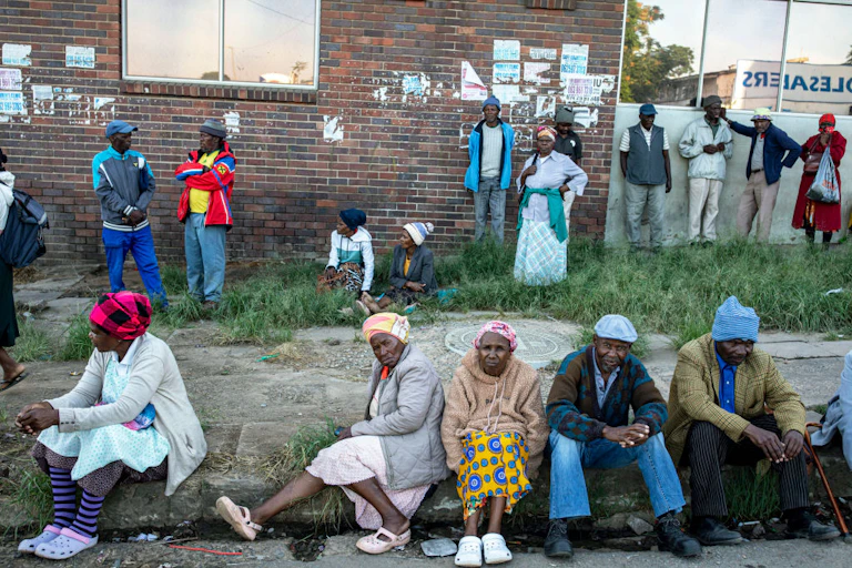 Older people sitting and standing on a pavement and grassy area next to a building