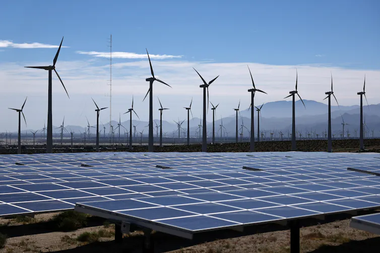 In the foreground, rows of solar panels point toward the sky. In the background, wind turbines spin.