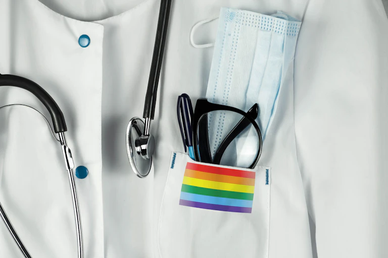 Close-up of rainbow flag sticker on the pocket of a doctor's white coat, also adorned with a stethoscope, glasses, pen and surgical mask
