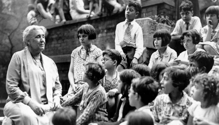 A black-and-white photograph shows a woman sitting outside, while rows of children sit on steps around her.