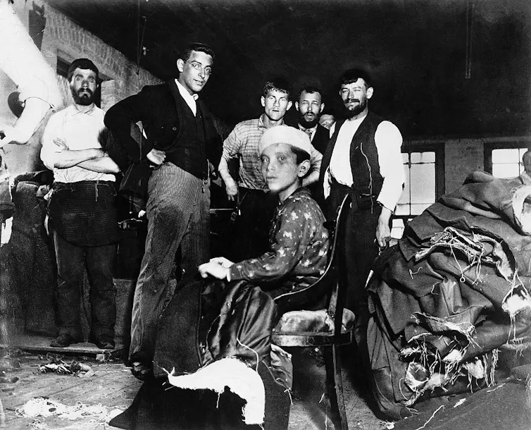 A black-and-white photo shows several standing men and a seated boy surrounded by piles of fabric.