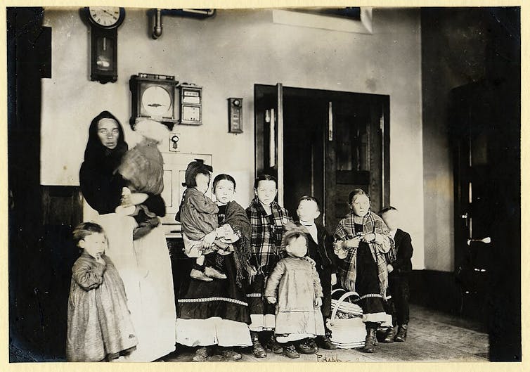 A black-and-white photograph of a woman in a dress and shawl, standing indoors with her young children, several of whom also wear dresses and shawls.