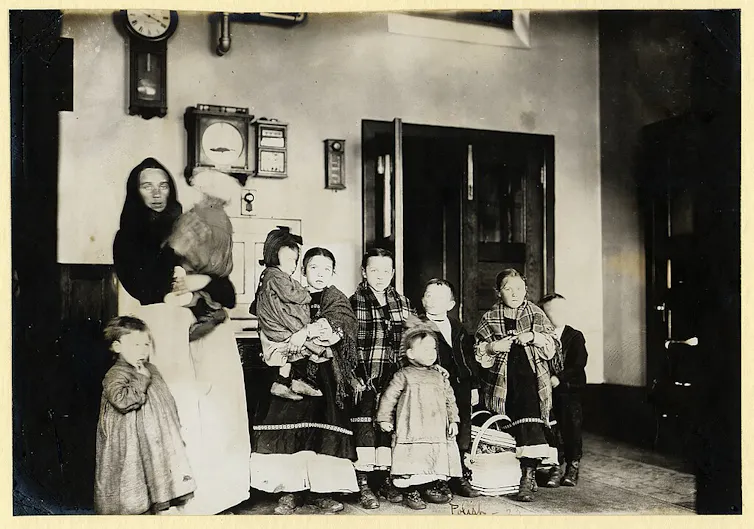 A black-and-white photograph of a woman in a dress and shawl, standing indoors with her young children, several of whom also wear dresses and shawls.