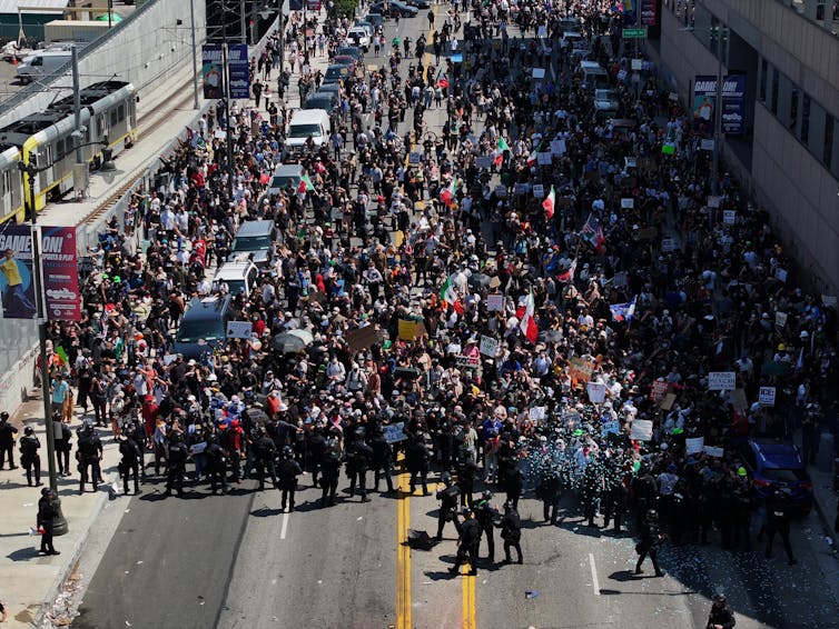 A large crowd of people stand together on a street, with a line of people in black standing close to them.