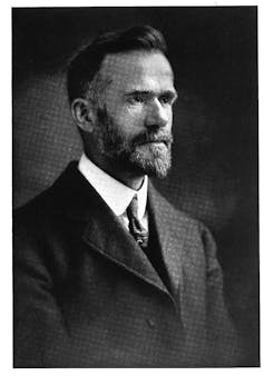 A black-and-white portrait of a man with a beard, looking serious, dressed in a suit and tie.