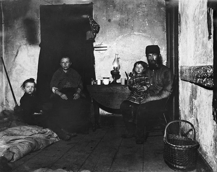 A black-and-white photo shows a man, woman and two children in a very simple room with dirty walls and almost no furniture.