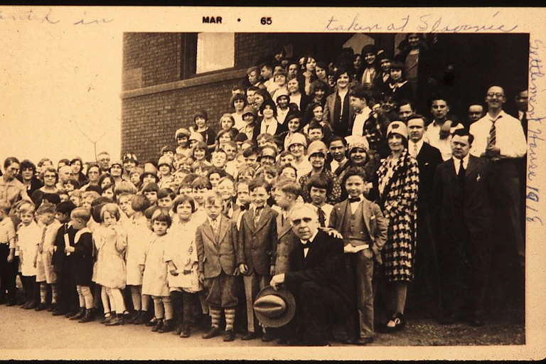 A sepia photo of rows of children and a handful of adults posing outside a brick building.
