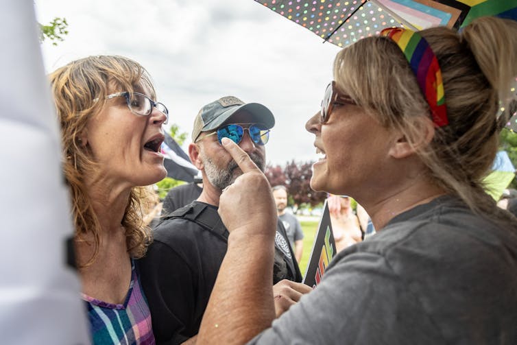 Two women arguing, watched over by a man in a hat.