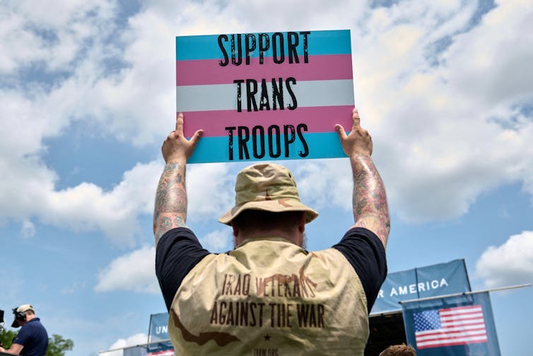 A person wearing a vest identifying him as a veteran holds a sign up that says 'SUPPORT TRANS TROOPS.'
