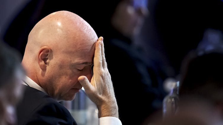 A bald man places his hand on his face.