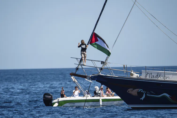 A young woman stands on the bow of a boat near a Palestinian flag.