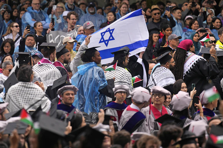A crowded scene of people in gray academic robes and caps, some of whom hold small Palestinian flags.