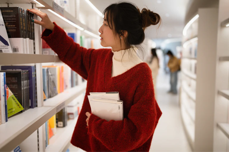 A woman wearing a red sweater selects books from shelves in a library.