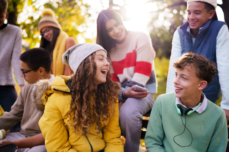 Happy teenagers laughing while sitting with their friends on a bench on an autumn day.