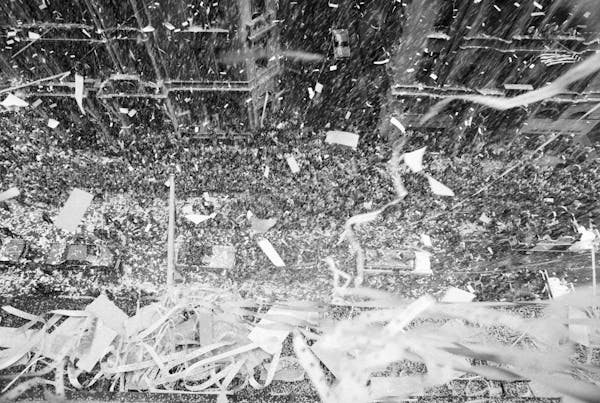 An avalanche of confetti rains down upon a cheering crowd and men in an open-topped car in a parade.