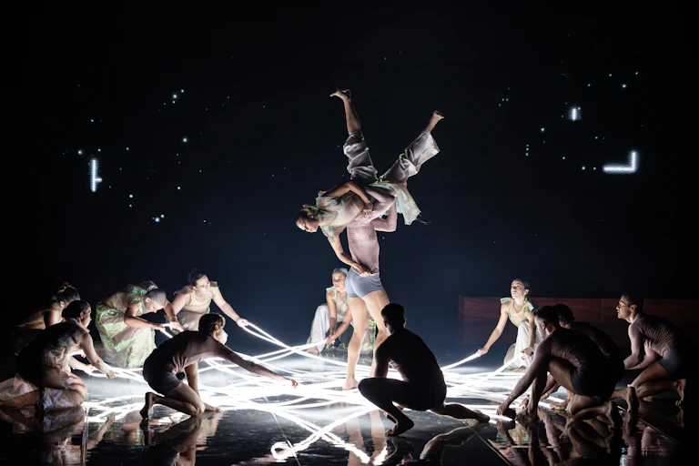 Production image: bodies hold strips of light on the floor, a woman is elevated upside down above a man's shoulders.