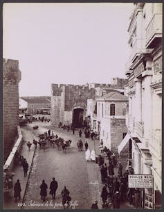 Picado de una calle en una ciudad con gente paseando y una puerta antigua al fondo.