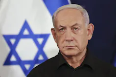 A balding grey-haired man looking stern with an Israeli flag behind him.