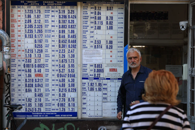 A man standing next to a board showing different currency exchange rates.