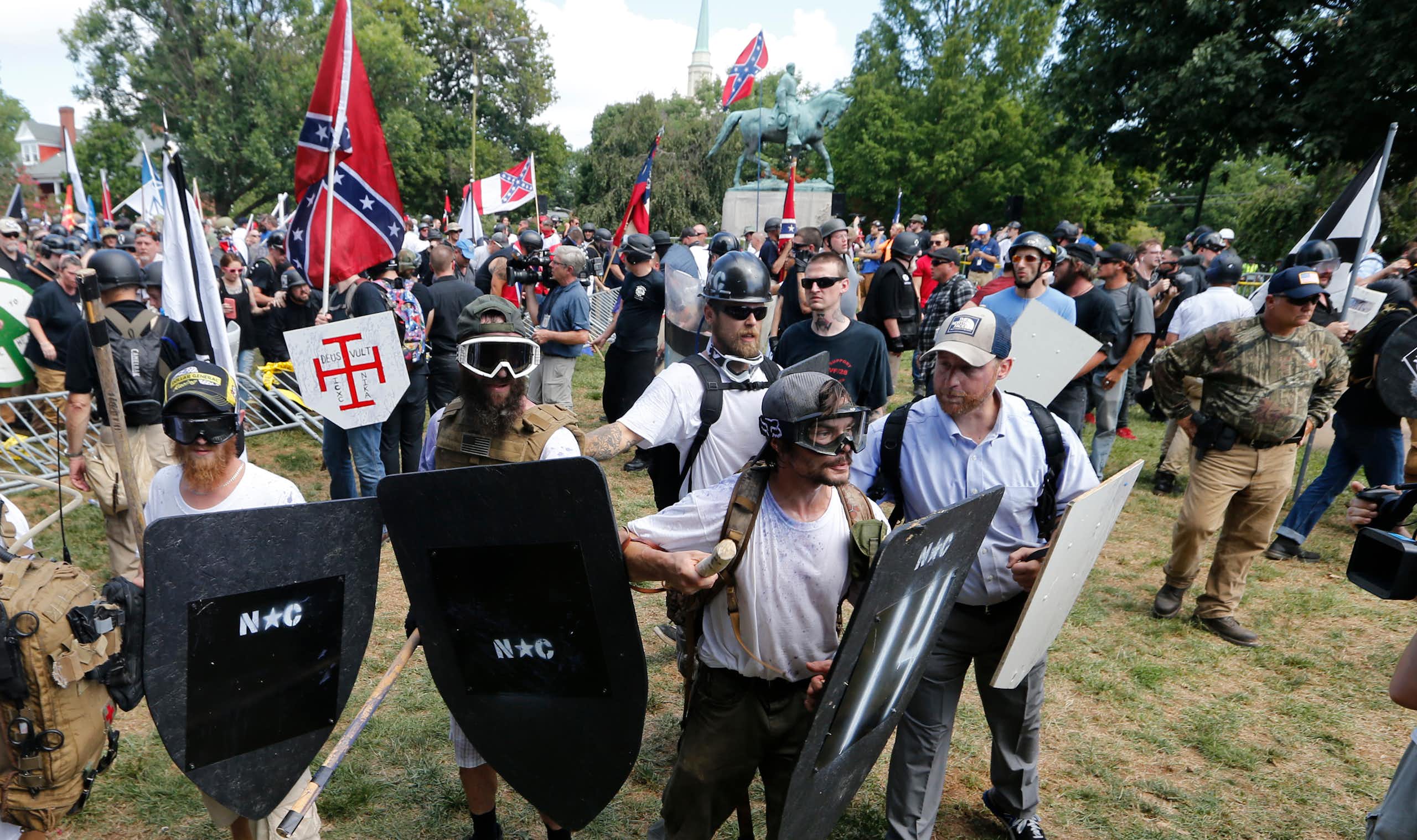 Men wearing helmets and holding shields stand near a monument of a man on horseback in a park.