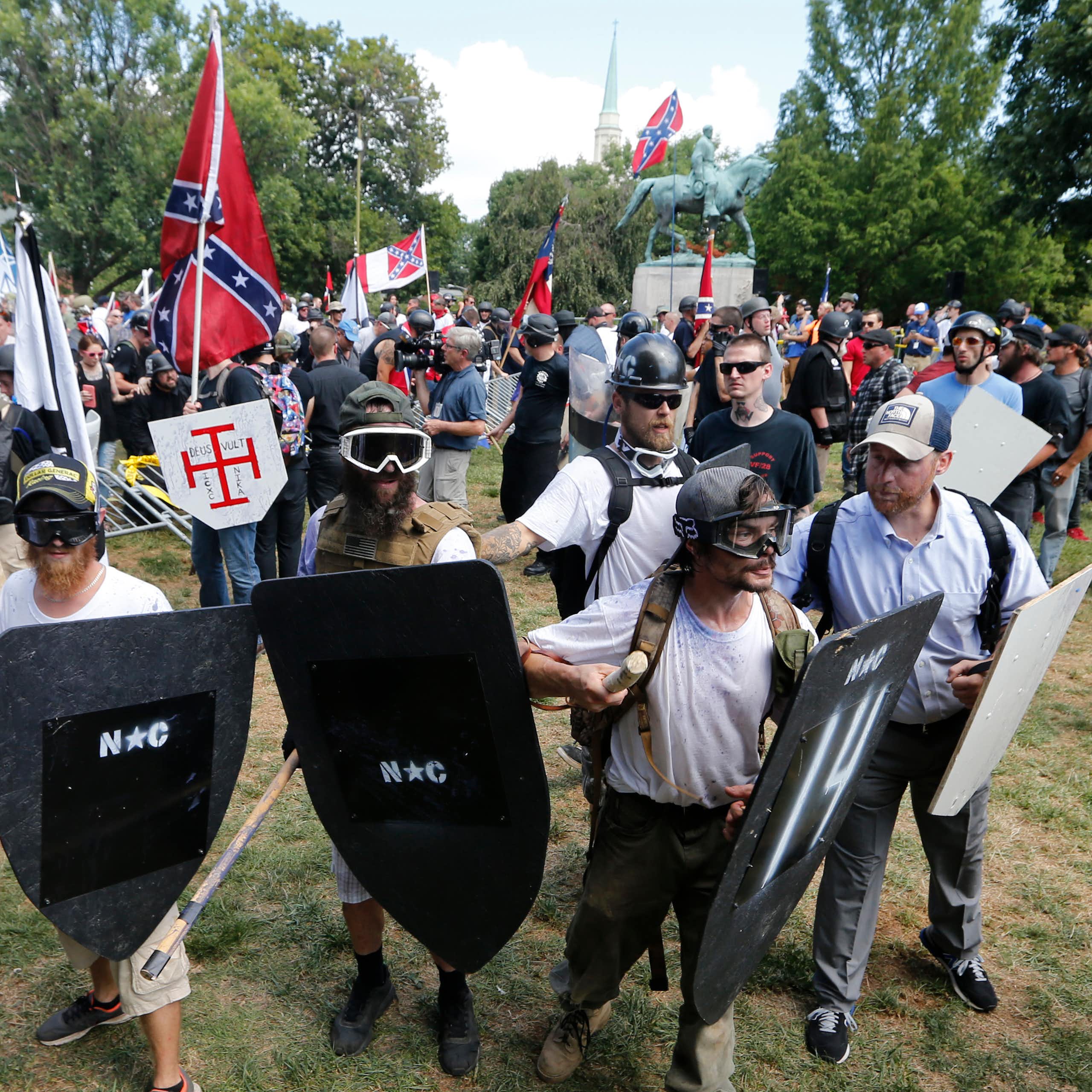 Men wearing helmets and holding shields stand near a monument of a man on horseback in a park.