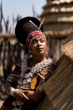 An African woman leans against a thatched structure wearing a high black headpiece fringed with red beads and shells around her neck.
