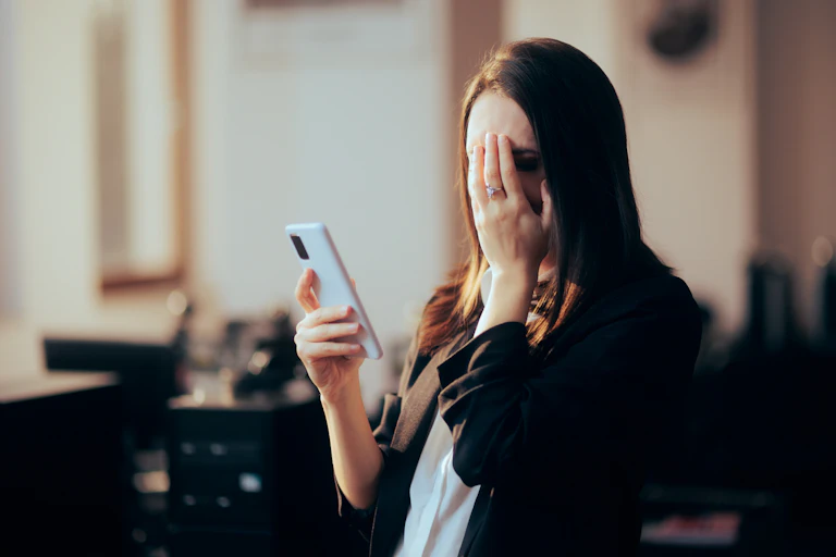 A woman covering her face with one hand, and holding her phone in the other