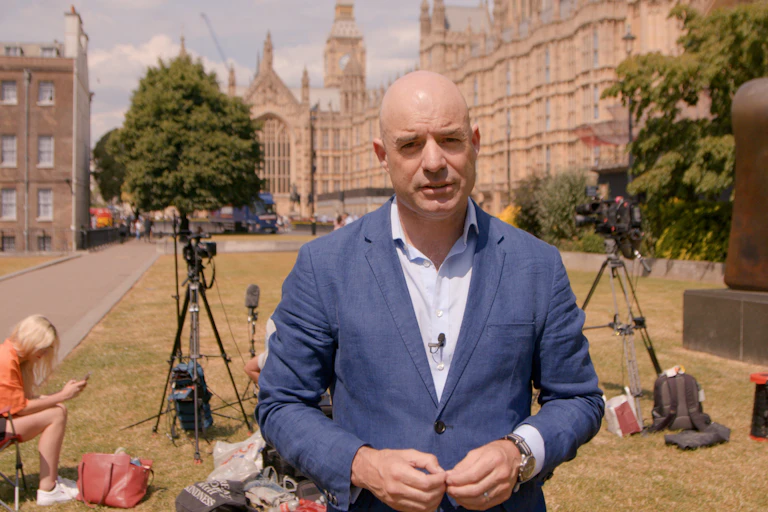 James Dyke stands by cameras talking outside on grass by Westminster