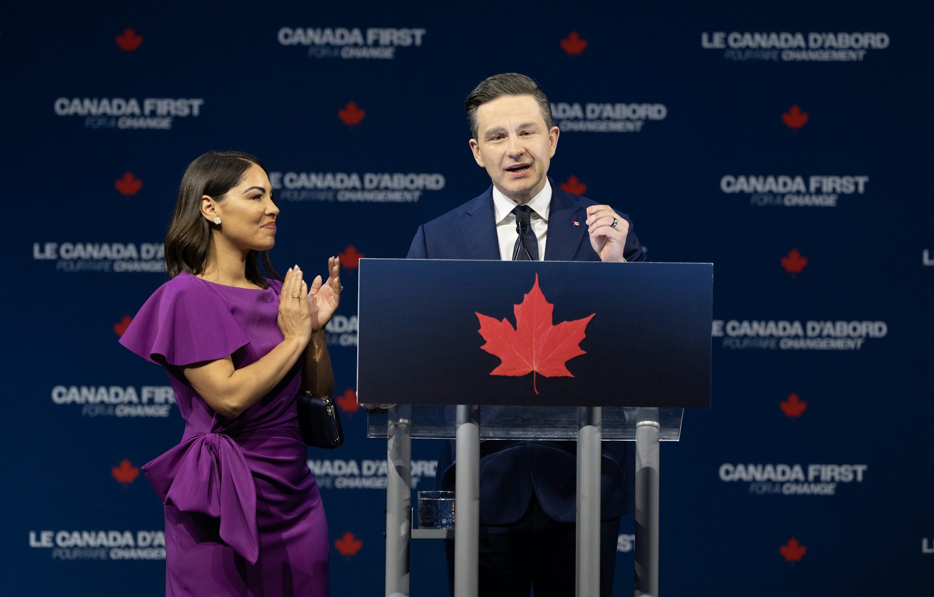 a woman claps and looks at a man in a suit who speaks at a podium gesturing with his hand