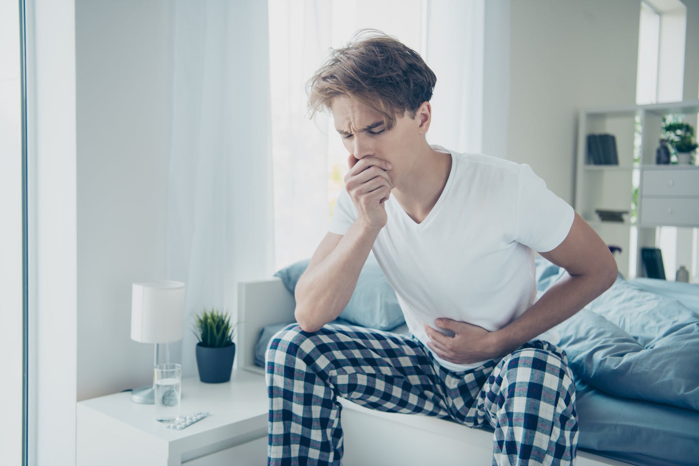 Stock photo of young man in a white T-shirt and plaid pajama pants sitting on the edge of a bed clutching his stomach with a hand over his mouth