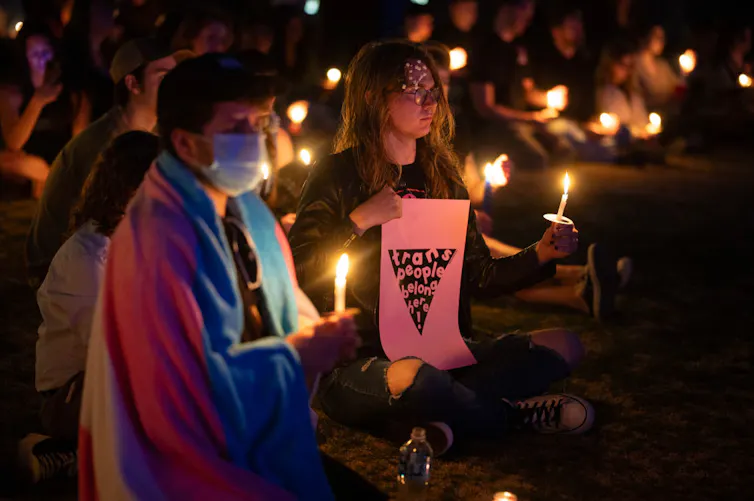 People sitting on the ground at night with lit candles in their hands. One person has a trans flag draped over their shoulders, another holds a sign reading 'trans people belong here!'