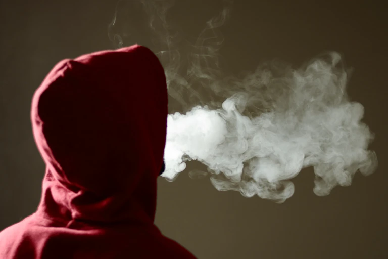 Young person facing backward in red hoodie exhaling a thick cloud of vaping smoke.