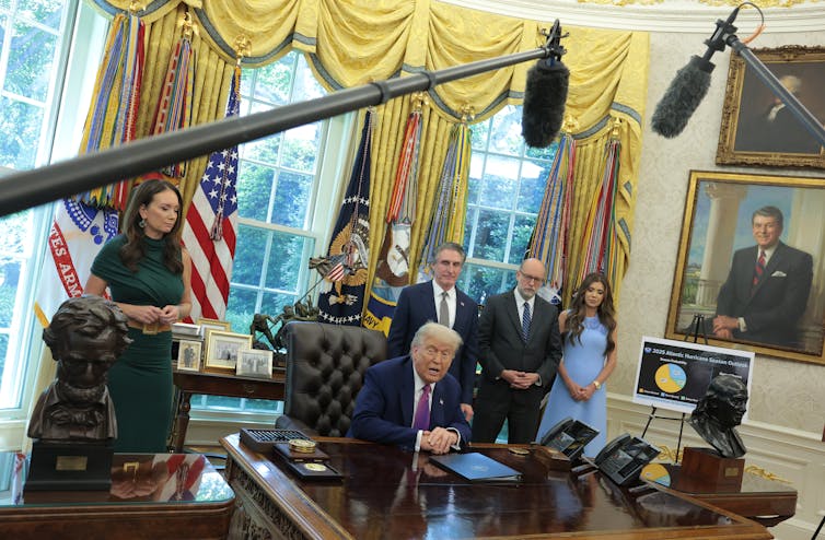 An older white man with a navy-blue suit sits at a large wooden table and is surrounded by four people dressed formally in an ornate room. Large microphones are seen hanging above the desk.