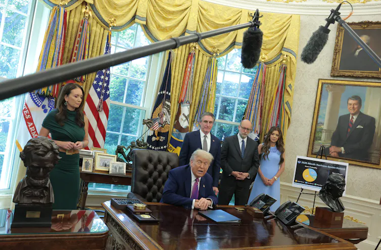 An older white man with a navy-blue suit sits at a large wooden table and is surrounded by four people dressed formally in an ornate room. Large microphones are seen hanging above the desk.