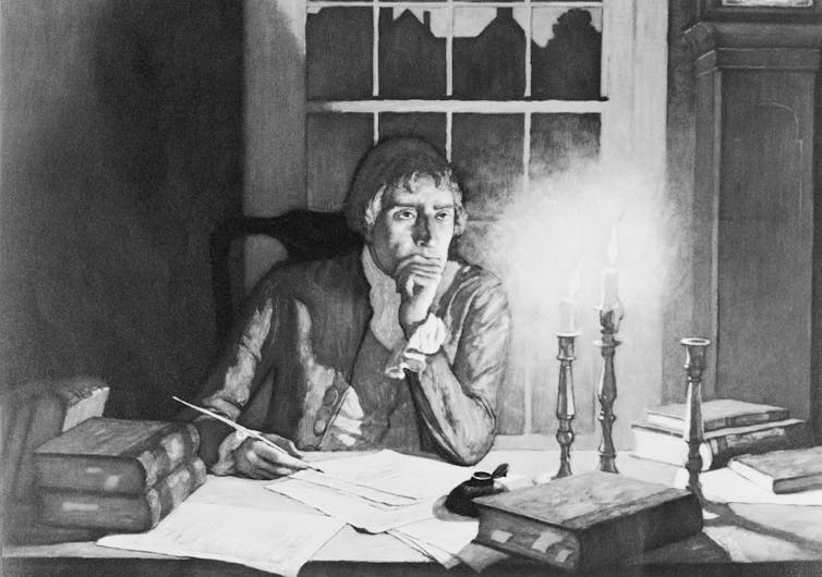 A man in colonial dress writing at a desk strewn with books and papers, by candlelight.