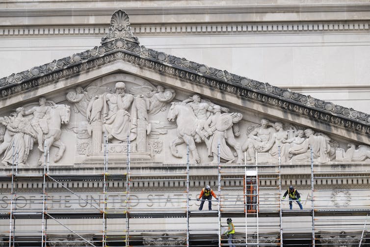 Workers stand on metal scaffolding near a very large, ornate marble building.