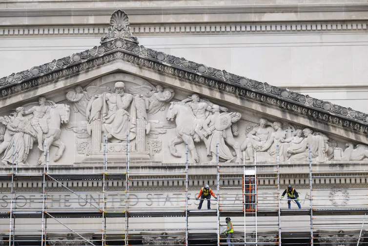 Workers stand on metal scaffolding near a very large, ornate marble building.