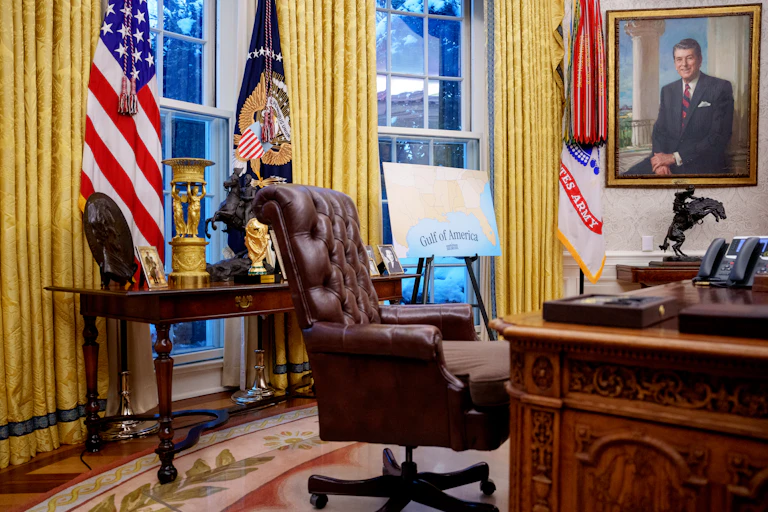 An empty desk is seen in a room with American flags, gold curtains and a map that says Gulf of America.