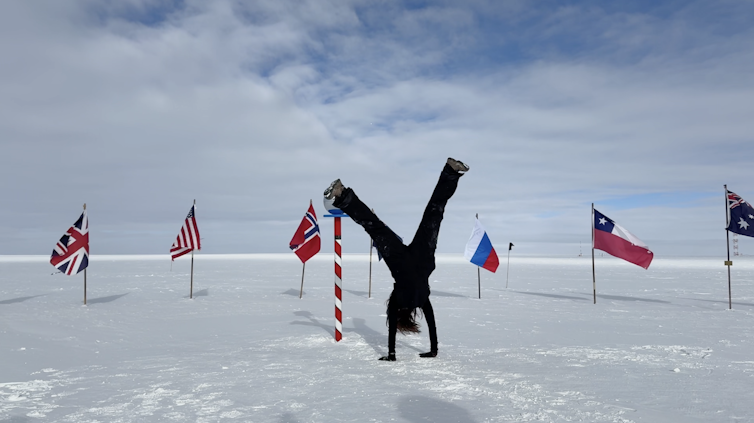 A person does a handstand on a white surface near a red-and-white striped pole surrounded by flags of various nations.