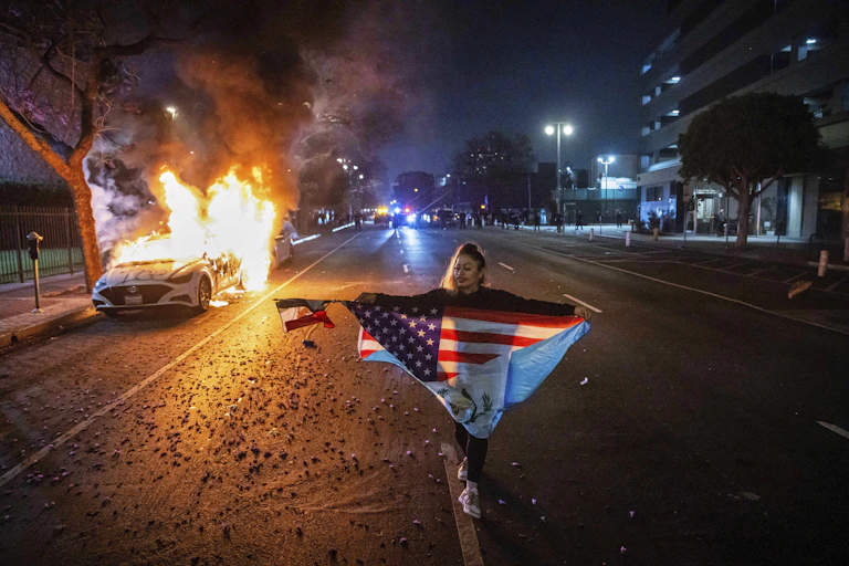 A person carrying multiple flags walks past a burning car during protests in Los Angeles.