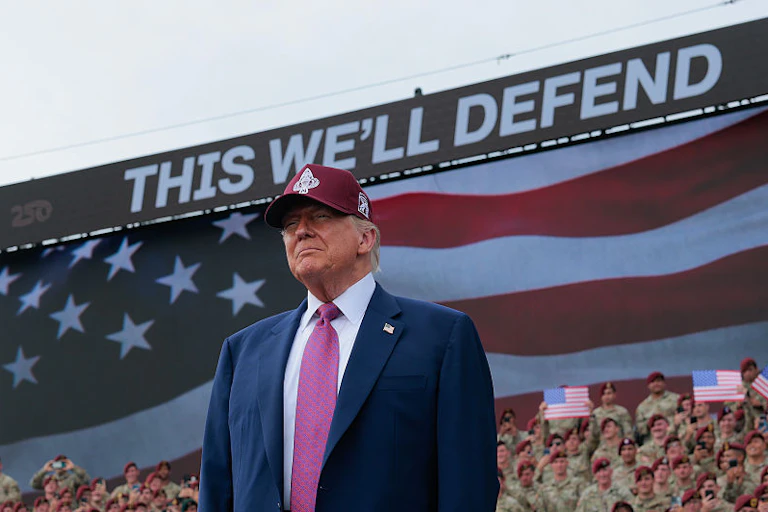 Man wearing a suit and cap standing in front of a crowd and a sign reading 'This we'll defend'