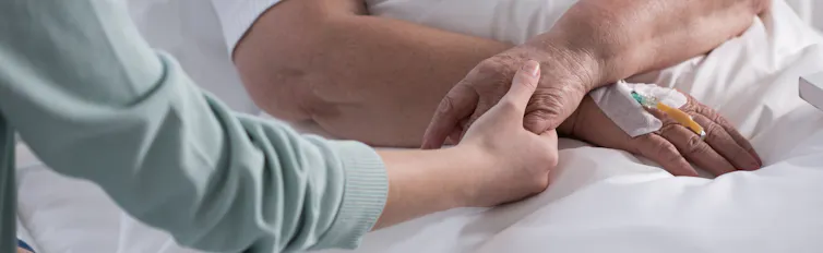 Cropped photo of an older person's hands, one with an intravenous tube, and one holding a younger woman's hand
