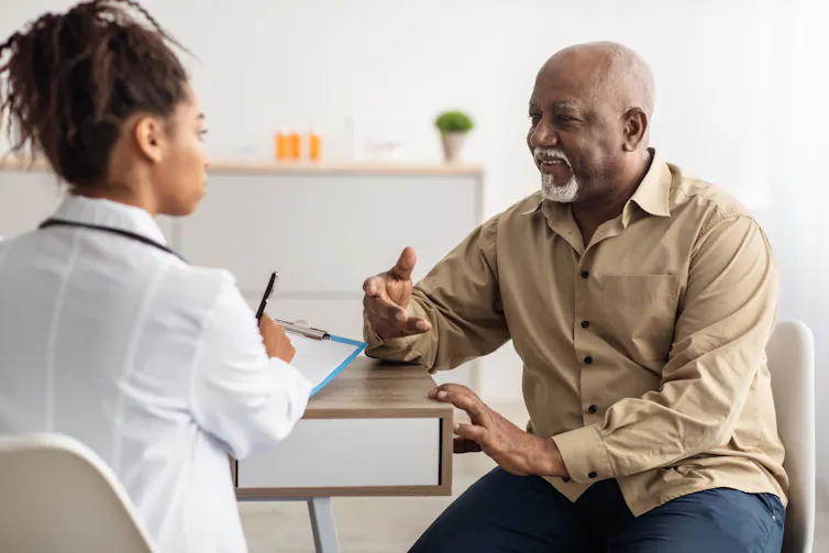 An older man with a white beard sitting at a table talking to a woman in a white coat with her back to the camera