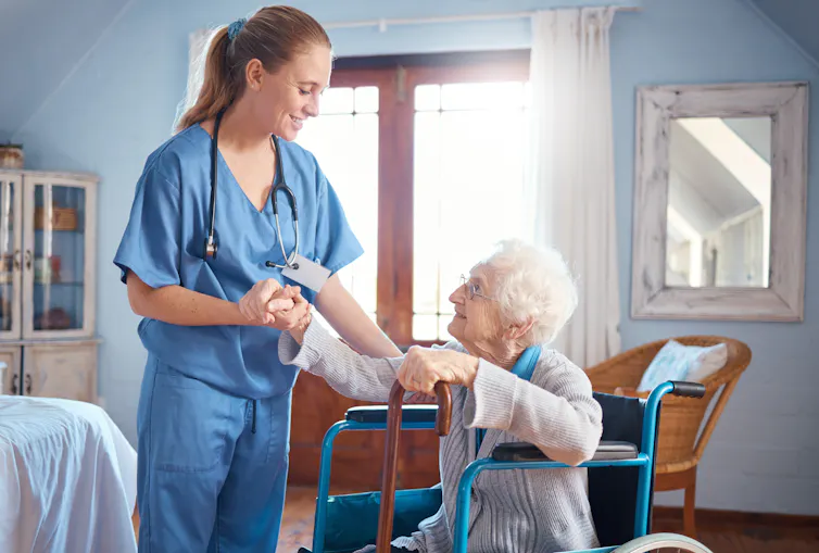 A woman with grey hair in a blue wheelchair, and a young woman in blue scrubs with a stethscope around her neck assisting her