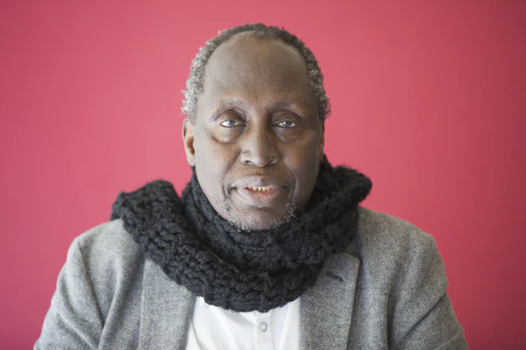 An older African man with a black scarf around his neck looks directly to camera against shocking pink background.