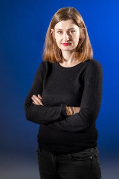 A blonde woman in a black sweater and dark jeans stands in front of a vibrant blue backdrop.