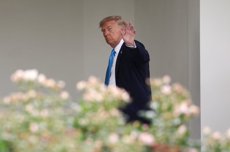 A man in a suit waving as he walks, behind flowers.