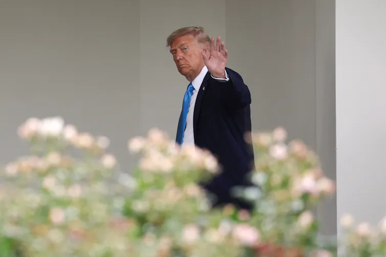 A man in a suit waving as he walks, behind flowers.