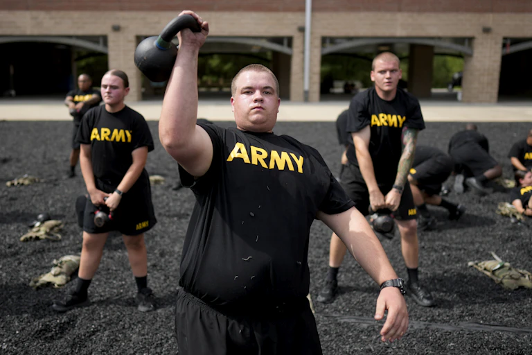 A man in black shorts and a black t-shirt lifts a kettlebell weights over his head, as three other people behind him hold weights.