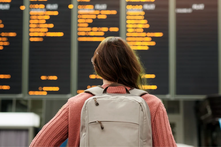 View from behind of a young woman in a backpack looking at a travel departures board